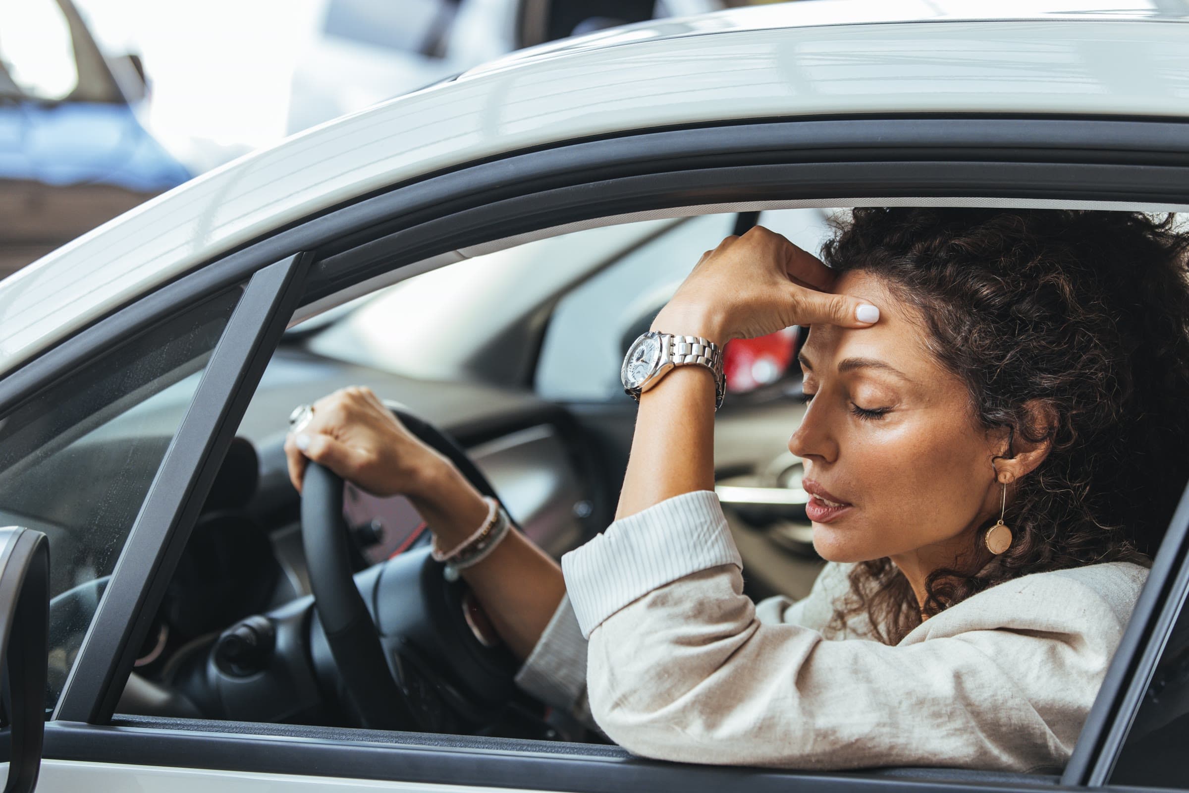 A woman in the driver's seat of a parked car, eyes closed, one hand on the wheel and the other pressed to her forehead.